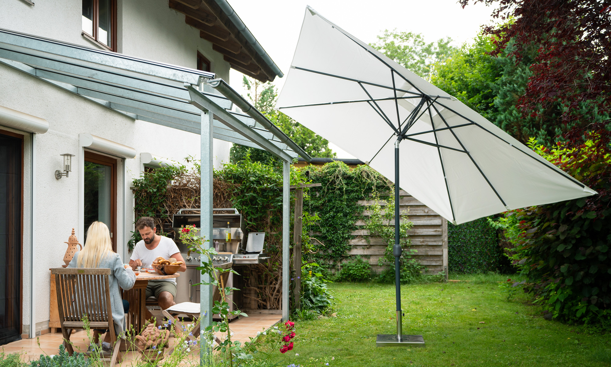Ombrellone con involucro a manovella in giardino accanto a terrazza e casa, persone sedute al tavolo